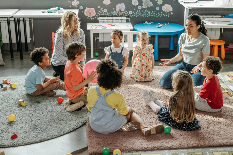 Group on children sitting on a floor surrounded by toys and two child care providers